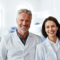 Two smiling dental professionals, a middle-aged man with grey hair and a young woman with brown hair, standing side by side in a bright dental office wearing white lab coats.